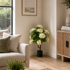 An artificial hydrangea bush with dense white flower clusters and green leaves, styled in a black pot on a light wood floor in a modern living room corner.
Real touch white hydrangea tree in black pot styled in a living room corner