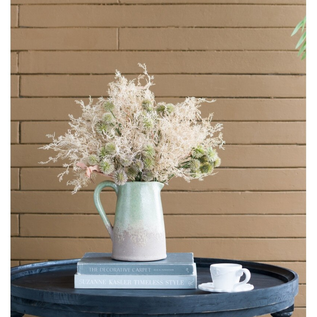 Thistle and Mist faux dried flower bouquet arranged in a green and cream ceramic jug on a dark round coffee table with styling books and a small white cup