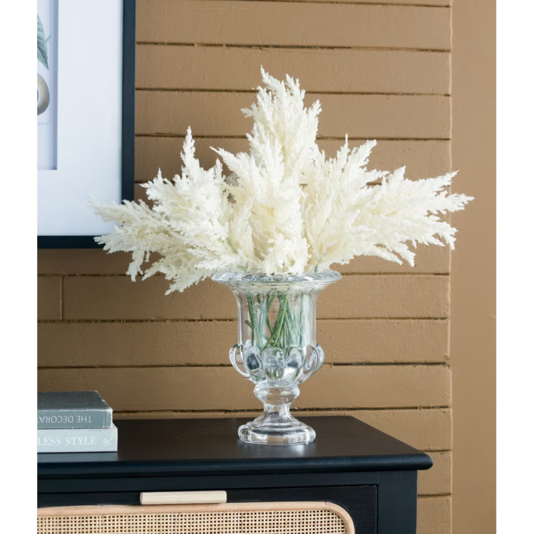 Multiple ivory faux pampas grass stems arranged in a clear glass urn vase on a dark sideboard, styled with framed botanical art and styling books against a terracotta brick wall