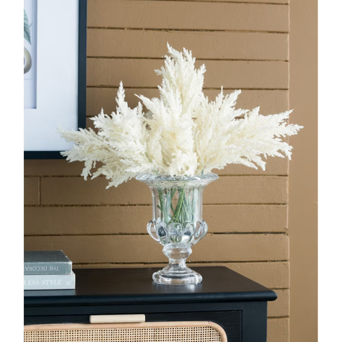 Multiple ivory faux pampas grass stems arranged in a clear glass urn vase on a dark sideboard, styled with framed botanical art and styling books against a terracotta brick wall