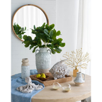 Small Aegean ceramic jug styled on a round wooden table alongside a large amphora vase with fiddle leaf branches, coral figurines, rattan tray and gold branch ornament, with a round timber mirror in the background