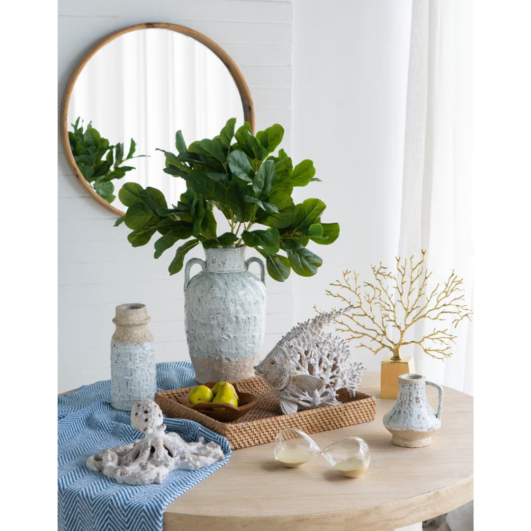 Small Aegean ceramic jug styled on a round wooden table alongside a large amphora vase with fiddle leaf branches, coral figurines, rattan tray and gold branch ornament, with a round timber mirror in the background