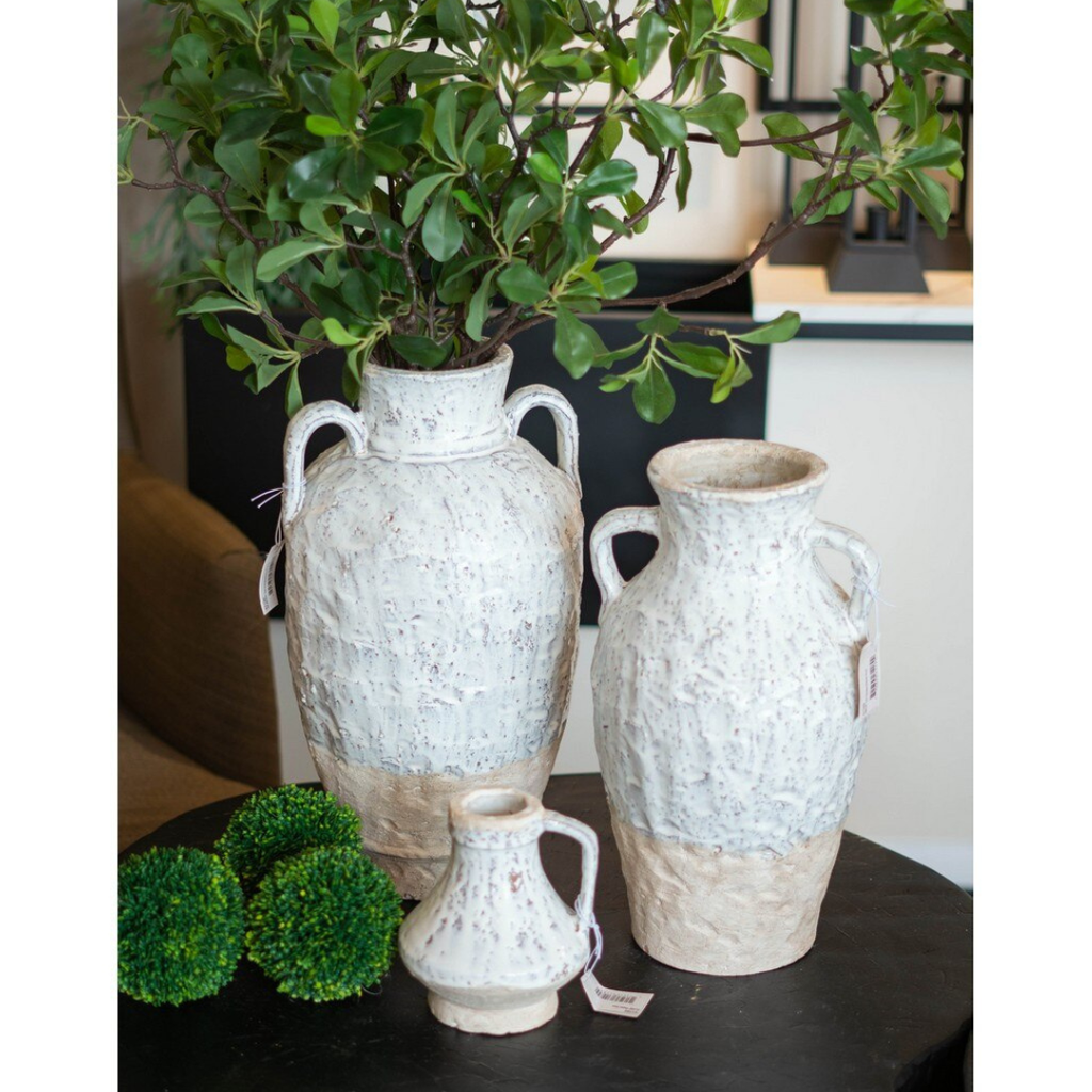 Three sizes of the Aegean ceramic collection grouped on a dark round coffee table — large double-handled amphora with greenery, medium single-handle jug, and small ceramic jug in the foreground with moss balls