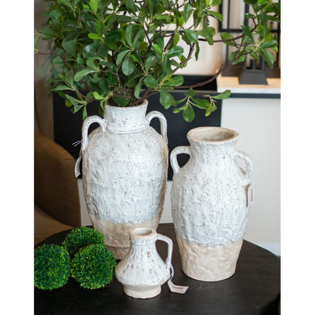 Three sizes of the Aegean ceramic collection grouped on a dark round coffee table — large double-handled amphora with greenery, medium single-handle jug, and small ceramic jug in the foreground with moss balls