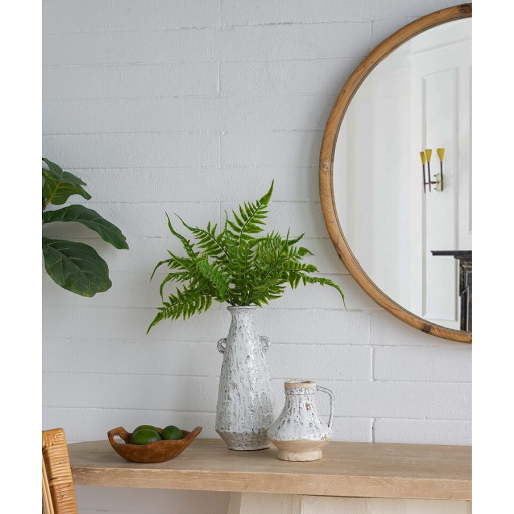 Aegean ceramic vase holding fresh fern fronds styled on a timber console table alongside the small Aegean ceramic jug, beneath a round timber-framed mirror against a white painted brick wall