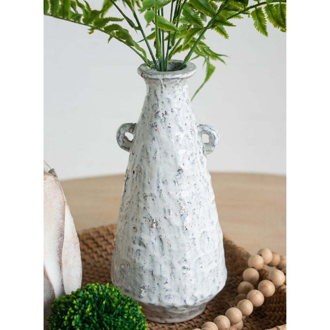 Close-up of Aegean ceramic vase holding fern fronds on a rattan surface, with moss ball and wooden bead garland in the foreground showing the heavily textured mottled glaze detail