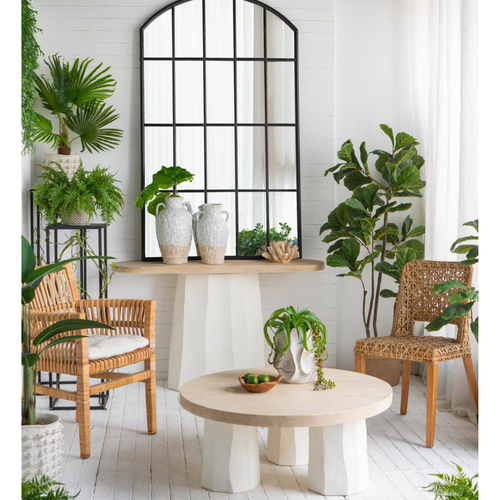 Two Aegean ceramic vases styled on a round timber pedestal dining table beneath a large arched black metal mirror, surrounded by fiddle leaf fig, monstera, palm and fern plants in a bright coastal boho room setting with rattan chairs
