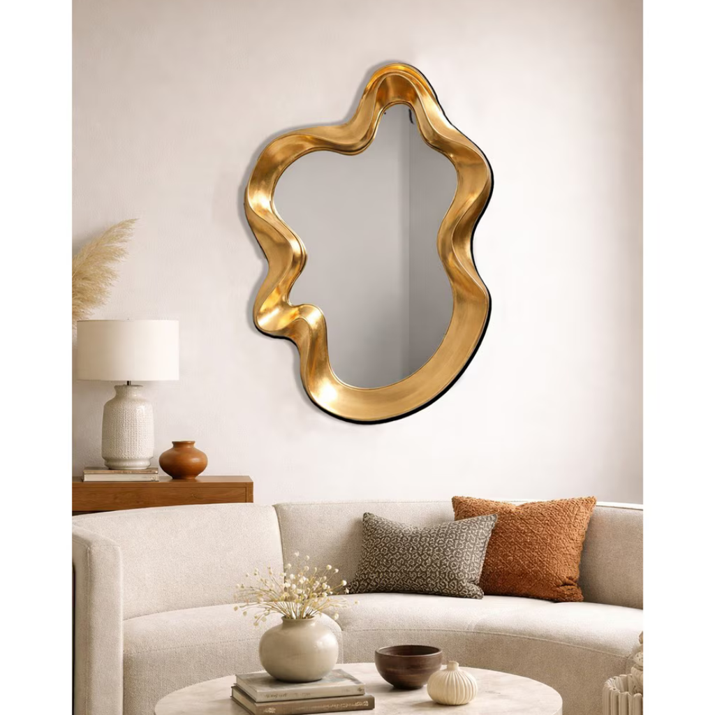 Gold sculptural wavy mirror hung above sofa in warm neutral living room styled with timber console and earthy cushions — Mish Lifestyle NZ