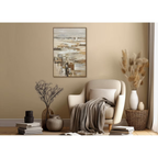 Cozy living room with a beige armchair, decorative plants, and abstract art on the wall. Abstract coastal seascape hand-painted canvas wall art with white sailboats on misty horizon in warm sandy neutrals and gold — framed original artwork in ornate dark bronze and gold frame